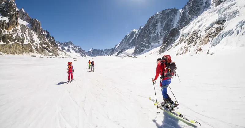 Vier skiërs met rugzakken doorkruisen een besneeuwd bergdal onder een strakblauwe hemel, omringd door hoge, ruige bergtoppen.